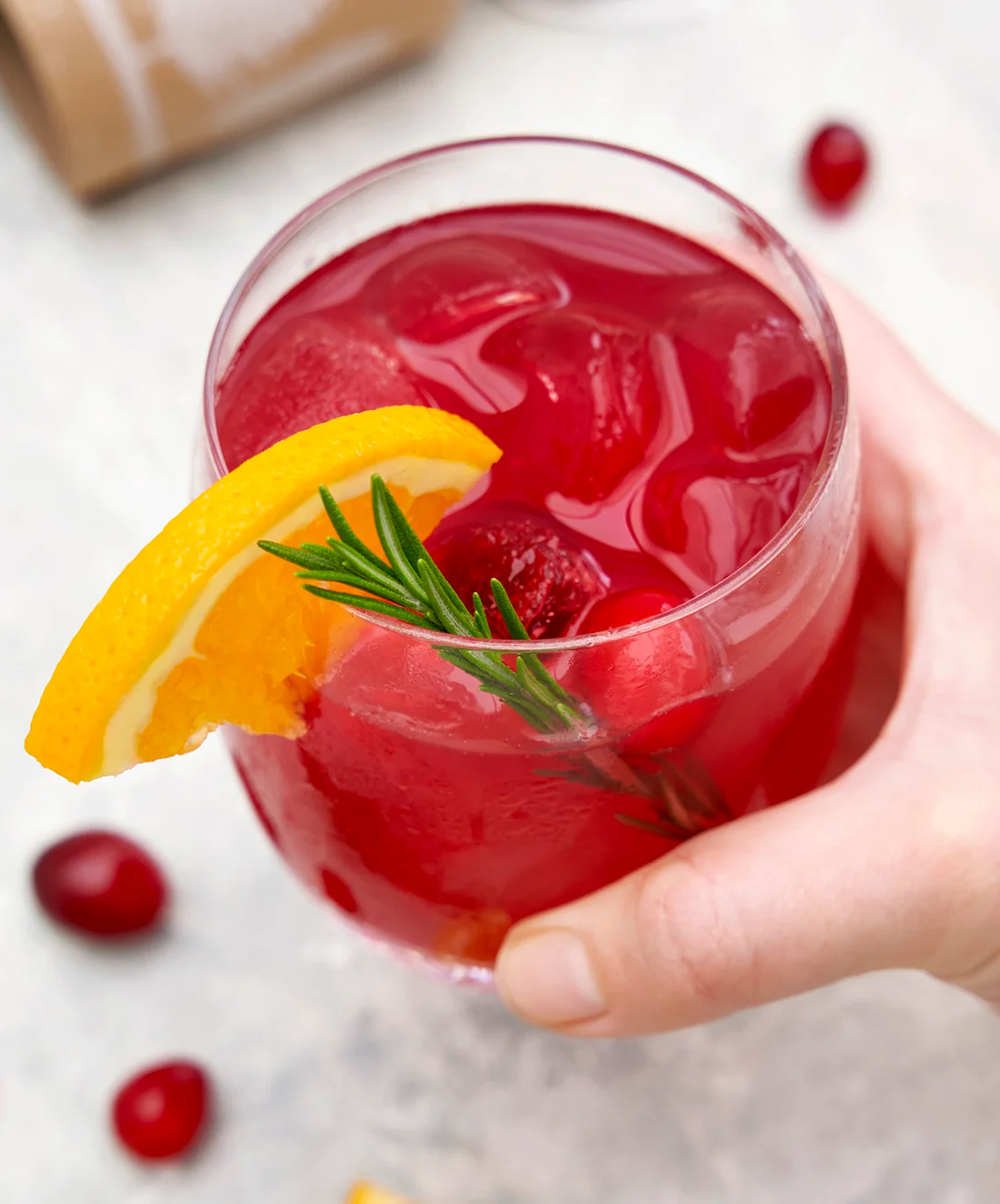 A hand holds a glass of bright red cocktail with ice, garnished with an orange slice and a rosemary sprig.
